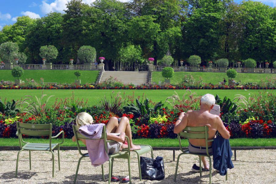 This is an image of the lovely greenery in Jardin du Luxembourg. There are two people sitting on the green chairs reading newspapers.