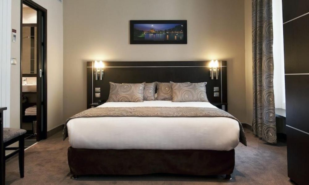This is an image of a darker hotel room with a queen bed and an ensuite, plus two lamp lights on the bed side that are on.