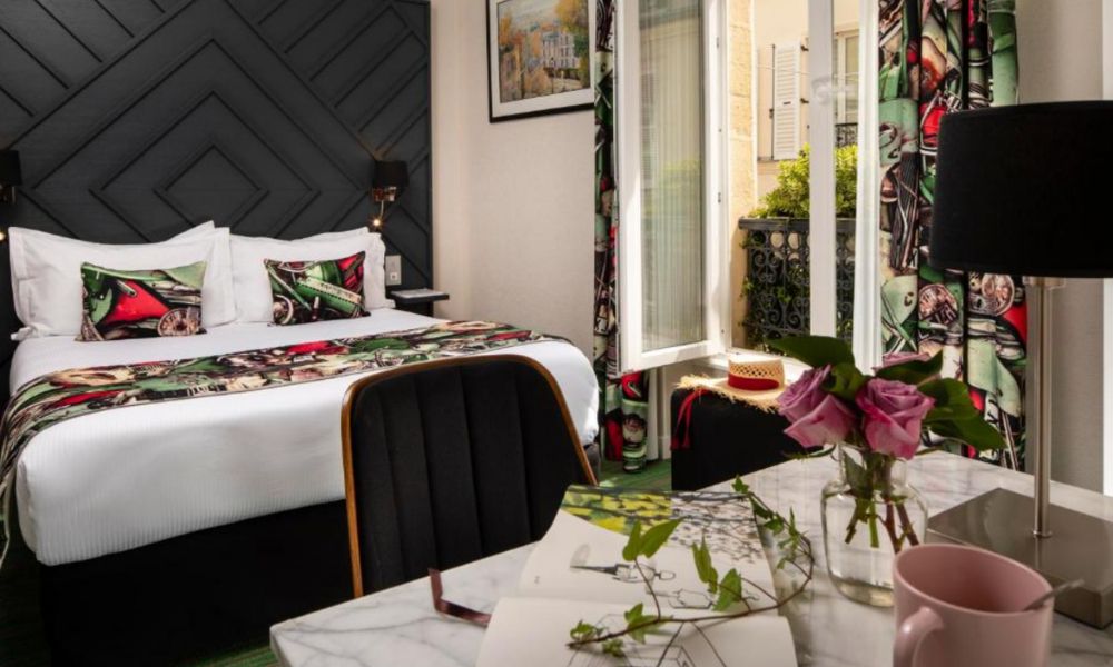 This is an image of a bedroom with artsy, floral patterns on the cushions, and blinds. There is also a big desk across from the double bed.