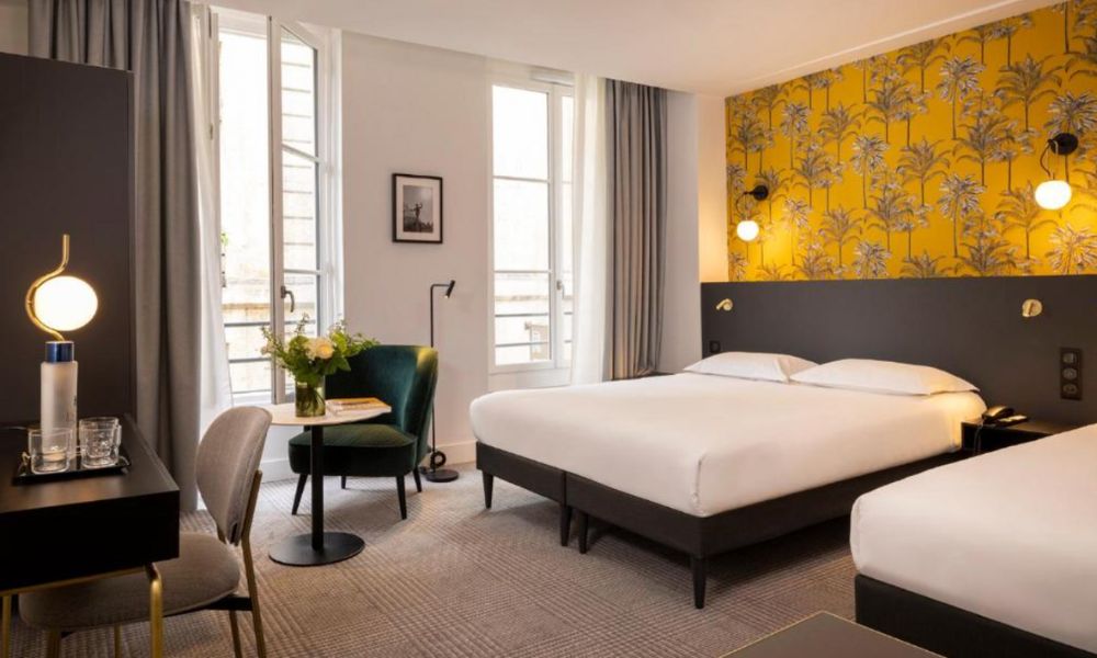 This is an image of a large hotel room with two beds, a coffee table by the window, a desk and a yellow-coloured feature wall.
