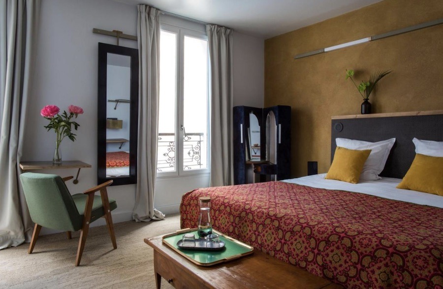 This is an image of a hotel room which is pretty minimalist other than its yellow feature wall. The bed is a double and topped with a red coloured duvet. There is a small desk in the corner with flowers on it.