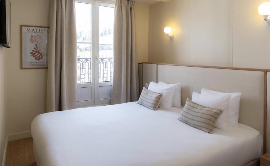 This is a picture of a minimalist hotel room with a double bed in the centre draped in white duvet covers. There is a small Matisse painting in the left corner hanging on the wall.
