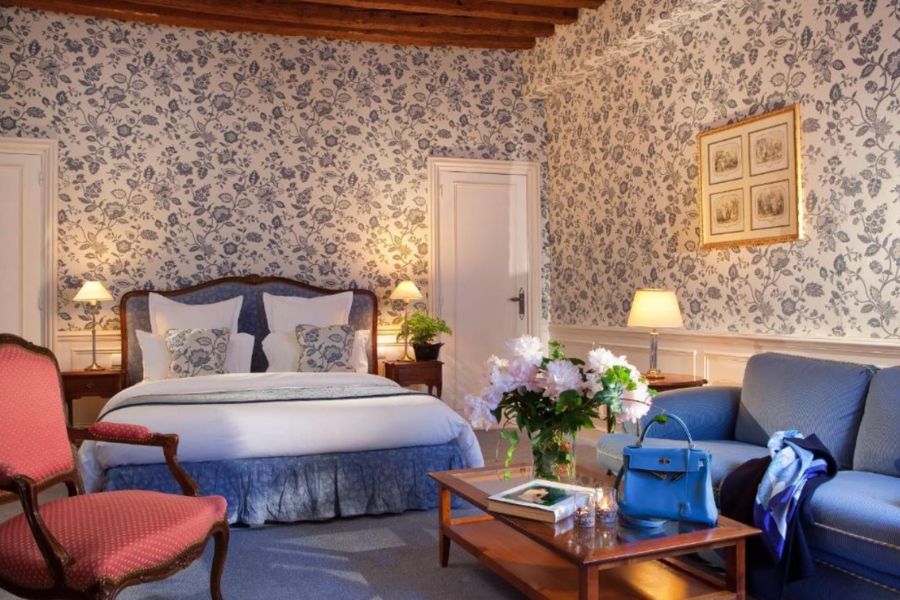 Elegant hotel room with blue floral wallpaper, exposed wooden ceiling beams, a Louis XV-style double bed, pink armchair, blue sofa, and fresh peonies on a coffee table