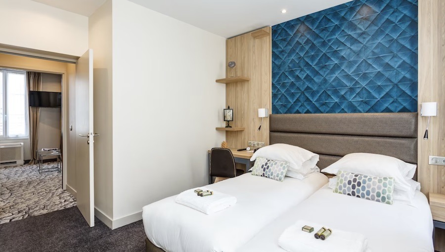 This is an image of a basic apart-hotel room in Paris with two single beds, and an extra room with a couch and a dining table.