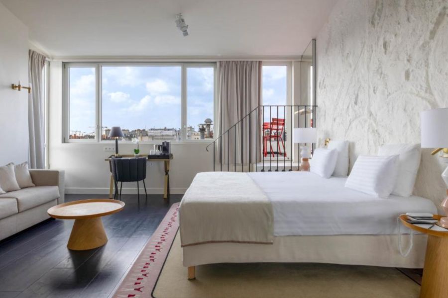 A bright Paris hotel room with white textured walls, dark wood floors, a king bed and a private balcony overlooking the rooftops
