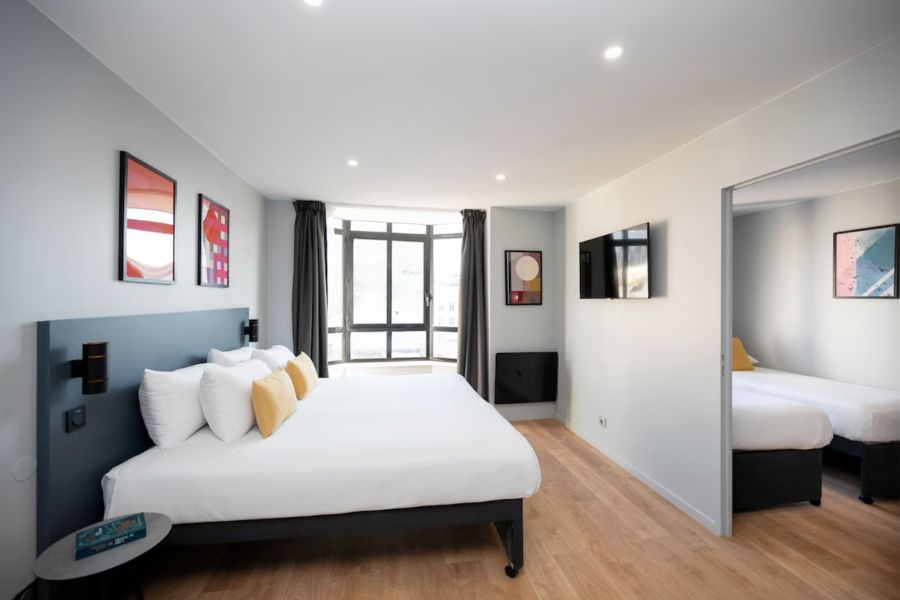 This is an image of a minimalist hotel room with a double bed. You can see another room on the side with two single beds in it. 