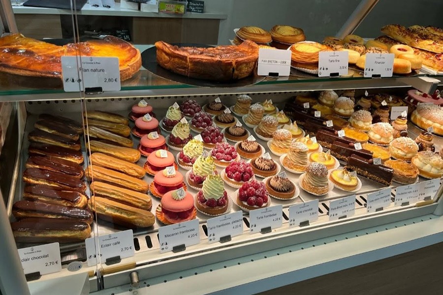 A bakery display case at Le Grenier à Pain filled with rows of pastries, including chocolate and coffee éclairs, raspberry macarons, fruit tarts, cream-topped choux buns, and flans, each with small price labels.