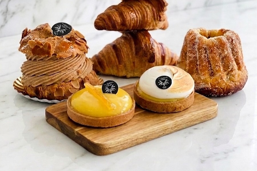 Assortment of pastries from La Maison d'Isabelle on a wooden board, including a stacked trio of croissants, a caramel-almond Paris-brest, a lemon tart, a meringue-topped tart, and a caneler, on a marble surface