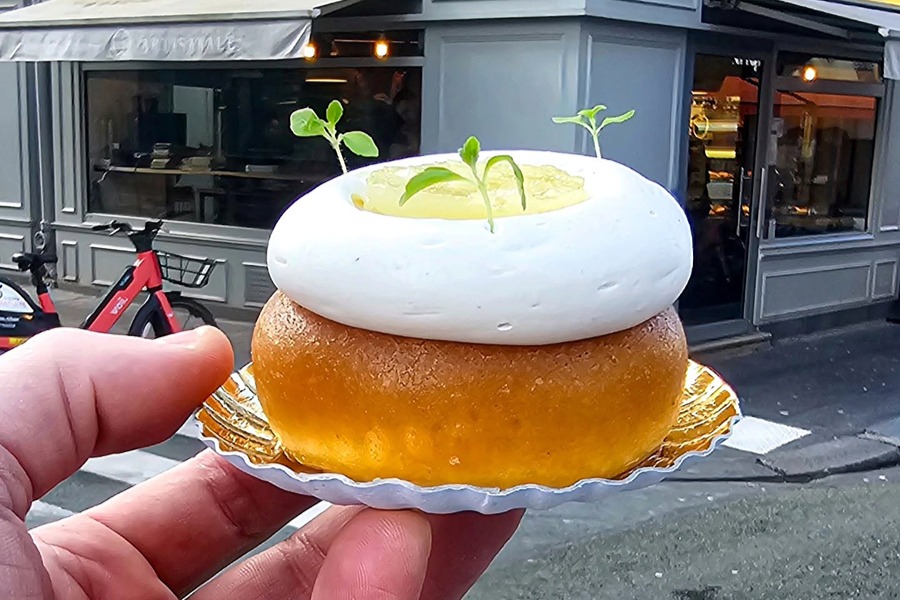 A hand holds a golden, glazed choux pastry topped with a swirl of white meringue-like cream, a pool of yellow curd, and small green sprigs, in front of a Boulangerie Utopia on a Paris street corner
