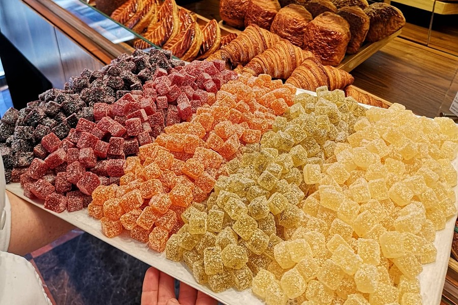 Tray of assorted sugared fruit jellies in a rainbow of colors held in the foreground, with croissants, pain au chocolat, and pastries displayed on shelves at des Gâteaux et du Pain bakery.