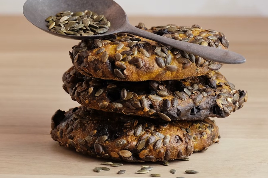 Stack of three seeded bread rolls generously coated in pumpkin seeds, with a rustic metal spoon of pumpkin seeds resting on top, on a wooden surface.