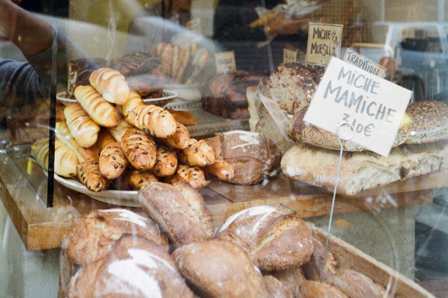 Bakery display window at Mamiche in Paris showing loaves of miche bread and chocolate chip rolls, with a handwritten 'Miche Mamiche' price sign, seen through reflective glass.