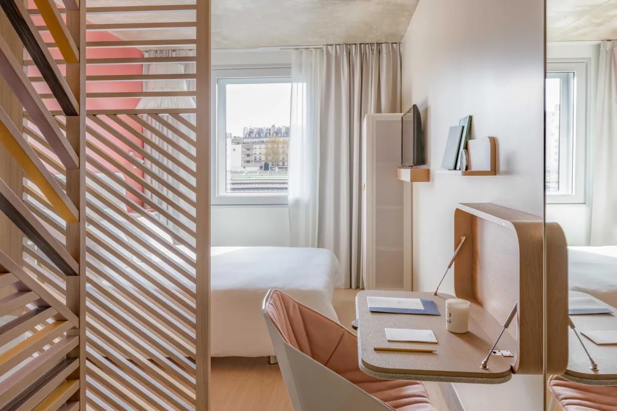 This is an image of a bright and dreamy hotel bedroom with a big double bed dressed with white sheets and subtle pink accents to the furniture.