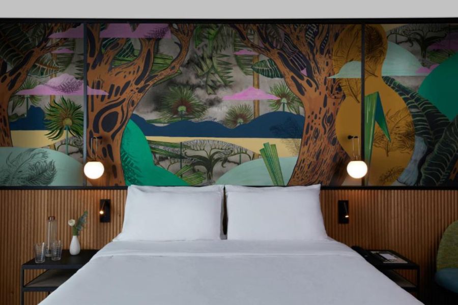 A hotel bed in Paris with white pillows against a bold illustrated mural of tropical trees and landscapes in greens, oranges and blues