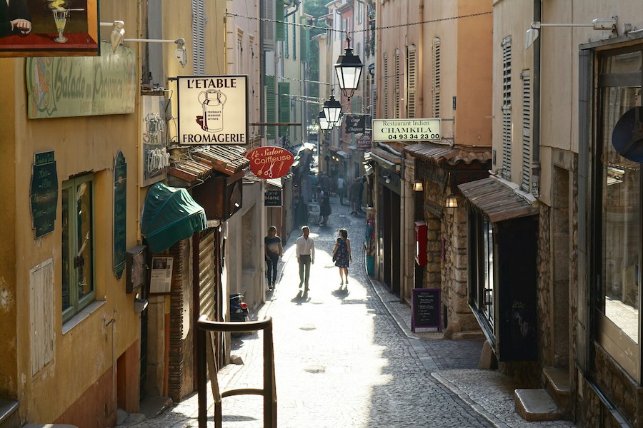 This is an image of a lovely local laneway in Paris. It is lined with shops and there are two people walking down it.