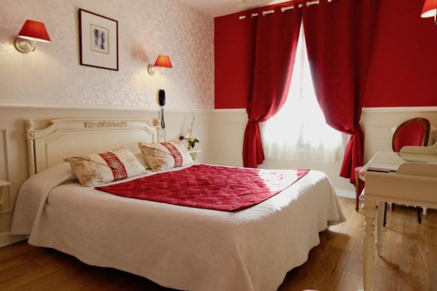 Classic Parisian hotel room with bold red curtains and accent wall, cream carved wooden headboard, red wall sconces, and a white quilted bedspread with red throw