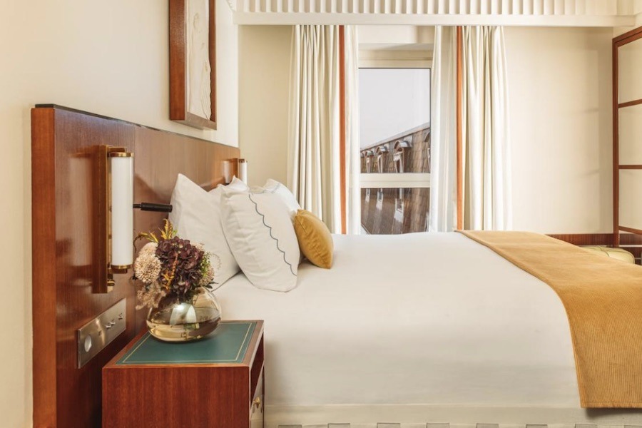 This is an image of a minimalist cream hotel room with a double bed, two side tables and a big window that's letting in lots of light.