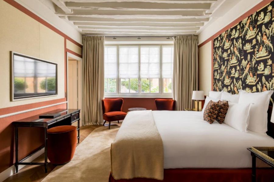 Contemporary hotel room with terracotta accents, exposed white ceiling beams, dramatic black chinoiserie wallpaper, rust-red velvet armchairs, and a large double bed