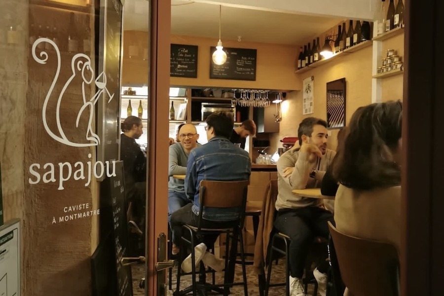 Interior of Sapajou, a cozy wine bar and caviste in Montmartre, Paris, with customers seated at high tables, bottle-lined shelves, and chalkboard menus on the walls