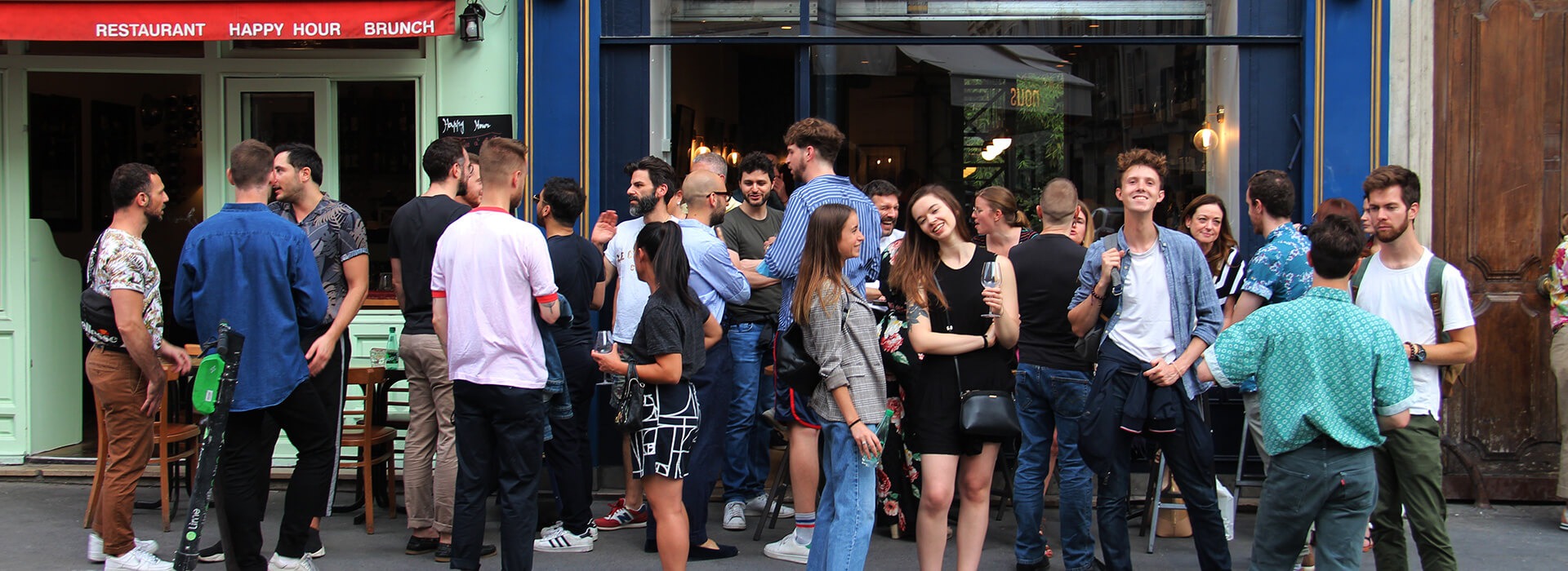 A large group of young people drinking wine in the street outside a wine bar.