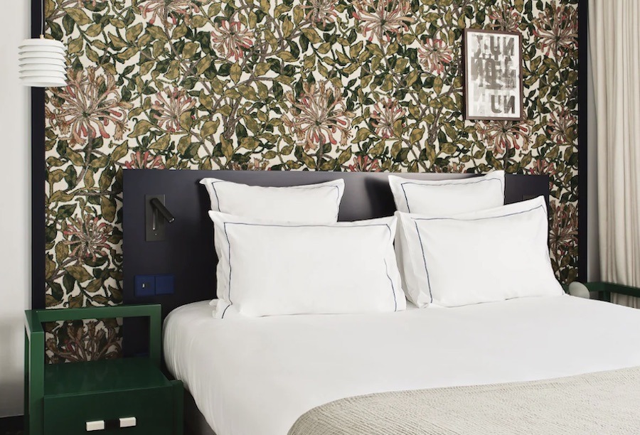 This is an image of a very nice hotel room with a double bed and a green bedside table. The feature wall behind it is full of flowers and greenery. 