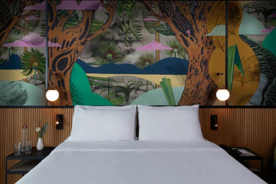 This is an image of an eccentric hotel room. There is a double bed with a white duvet and the wall behind it has feature art of trees and clouds. 