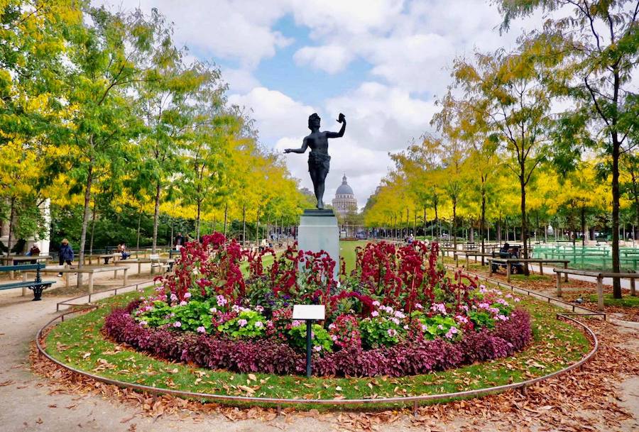 This is an image of Luxembourg Gardens main flower circle with a statue in the centre. You can see that trees line the rest of the path and they are turning yellow as the weather changes.