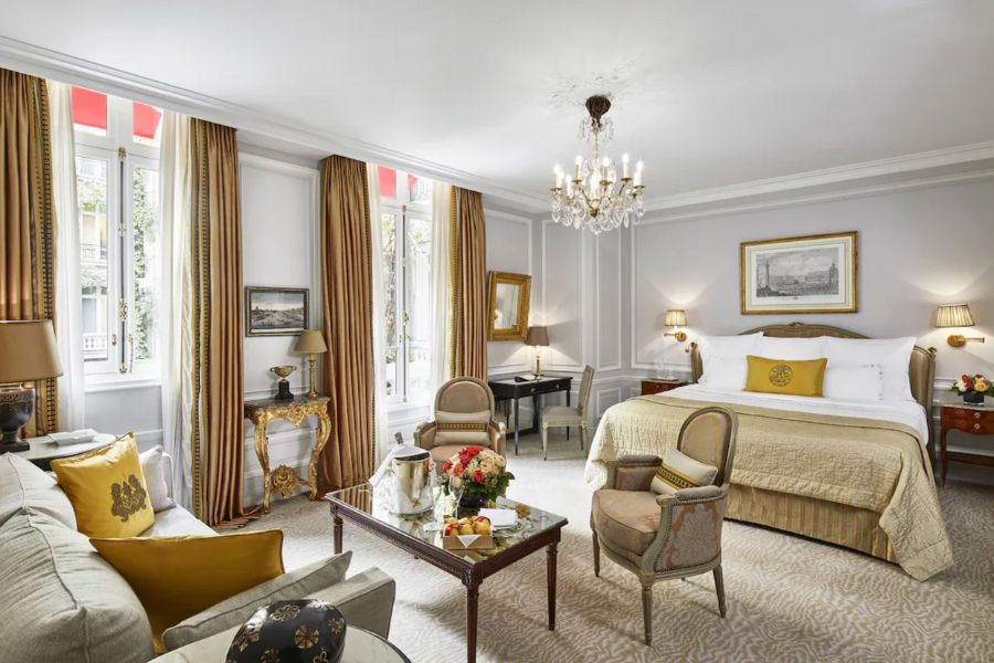 This is an image of a fancy hotel room in Paris with a big chandelier and a sitting area alongside the bed.