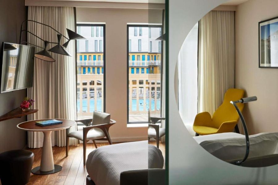 A hotel room in Paris with floor-to-ceiling windows overlooking an indoor swimming pool, warm neutral tones and a yellow accent chair