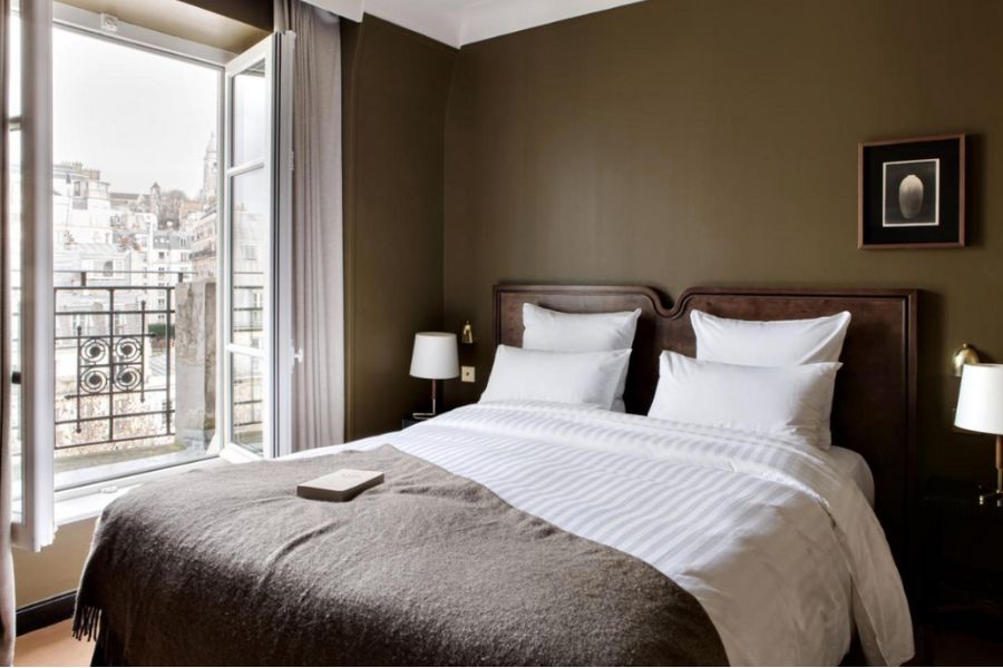 This is an image of a luxe hotel room. There is a double bed with a white duvet and brown throw rug. The rug matches the green/brown shade of the walls. 