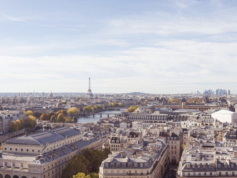 This is an image of Paris from a higher ground. You can see a skyline of buildings, bridges, the Seine, the Eiffel Tower and lots of trees that are turning yellow/orange.