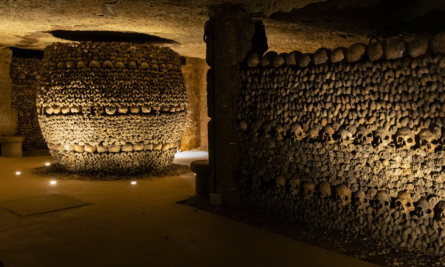 A dimly lit chamber in the Paris Catacombs featuring a round column and wall made from stacked skulls and bones, illuminated by soft ground lighting.