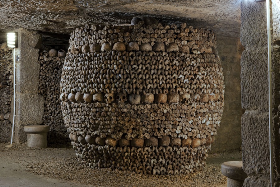 A large barrel-shaped column built from stacked bones and skulls in the Paris Catacombs, lit by a wall-mounted light.