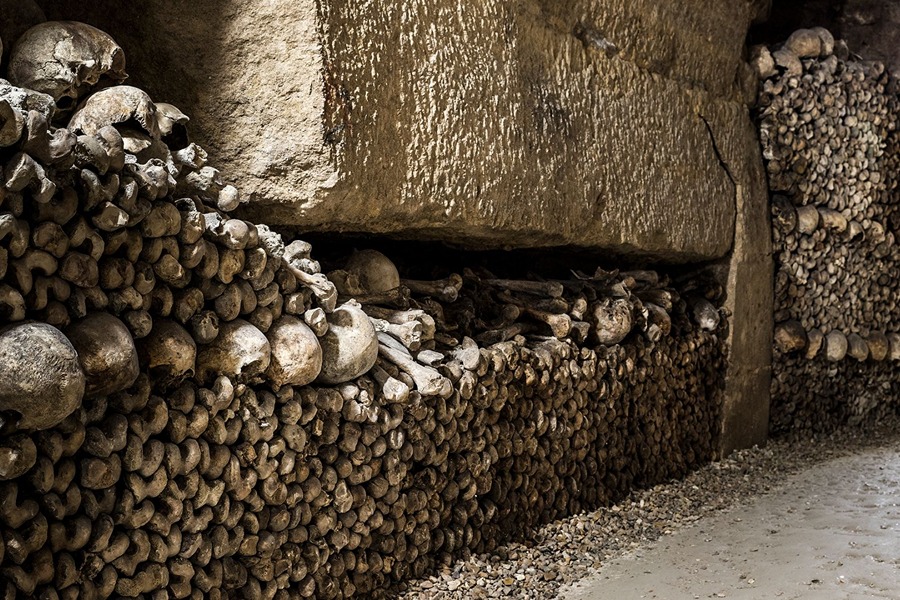 Rows of stacked skulls and bones lining the base of a stone wall along a Paris Catacombs corridor.