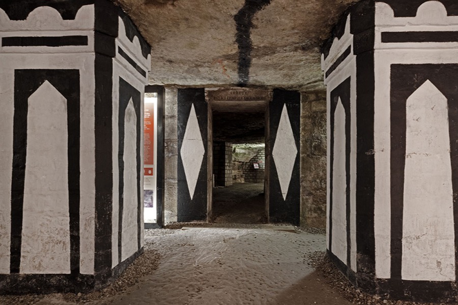 A stone archway in the Paris Catacombs flanked by black-and-white painted pillars, leading into a dimly lit tunnel.