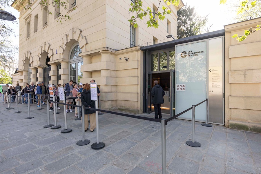 Visitors queuing outside the entrance to Les Catacombes de Paris, with roped stanchions lining the sidewalk beside the building.