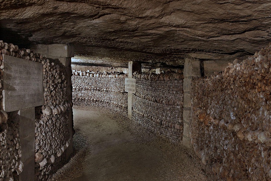 A long ossuary corridor in the Paris Catacombs with walls of stacked bones and skulls, an engraved plaque, and a stone cross marker.