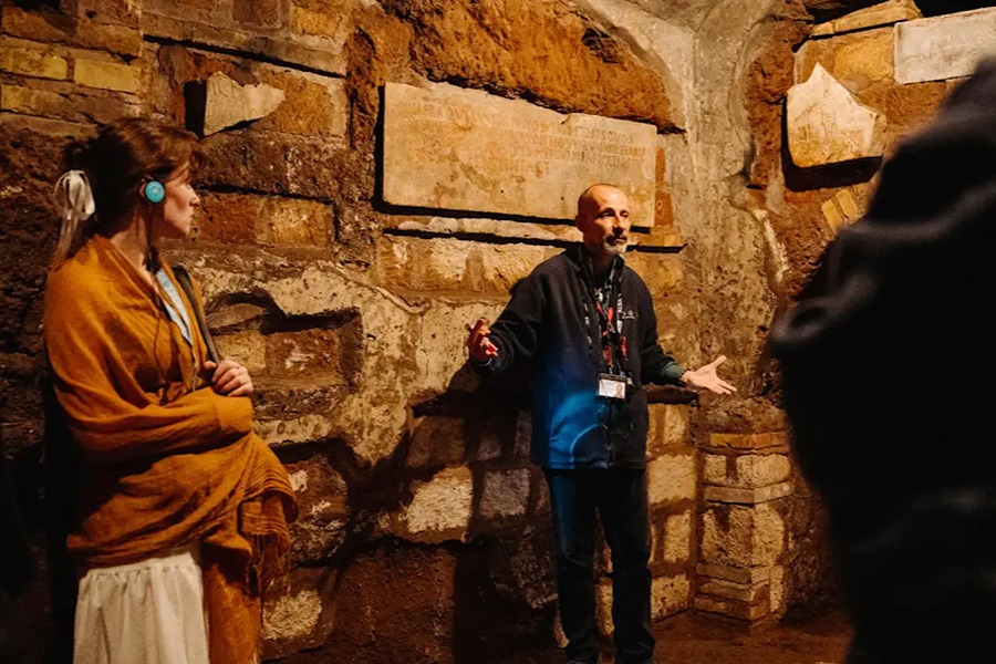 A tour guide explains an engraved stone inscription to visitors, one wearing an audio headset, inside the Paris Catacombs.