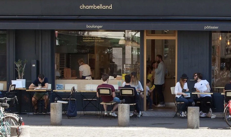 This is an image of a shop-front in Paris with lots of seats out front with people eating and drinking coffee.