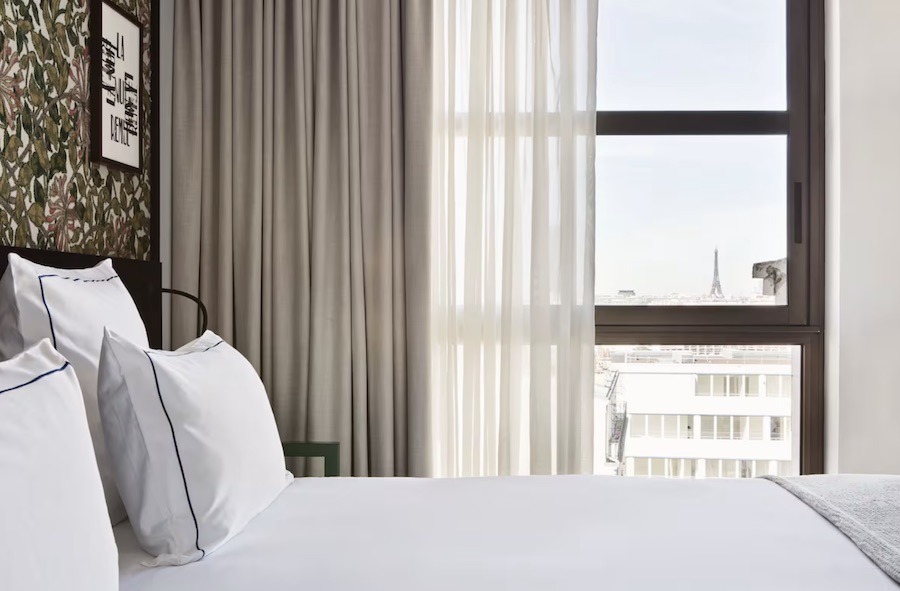 This is an image of a minimalist hotel room with a nice bright window that offers a view of the Eiffel Tower.