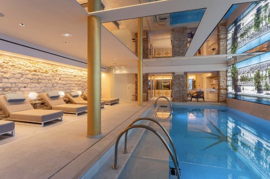 An indoor hotel pool in Paris with blue water, gold columns, exposed stone walls and a row of cream loungers alongside