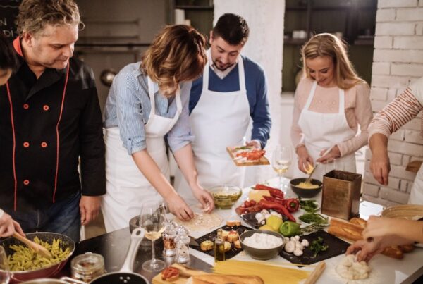 This is an image of a tour group with aprons on learning how to cook in front of a chef around a bench full of food.