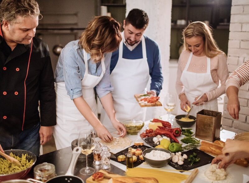 This is an image of a tour group with aprons on learning how to cook in front of a chef around a bench full of food.