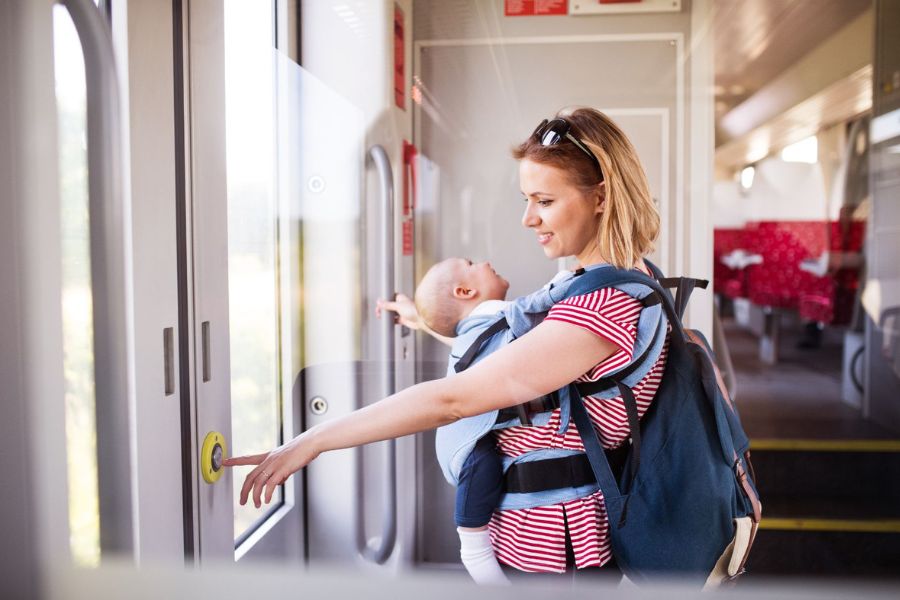 This is a picture of a mother on a train pressing the button to open the doors. She has a baby sling with a young child in front of her.
