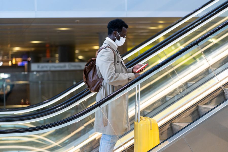 This is an image of a man standing to the right on an escalator. He is looking down at his phone and is holding on to a small carry-on suitcase.