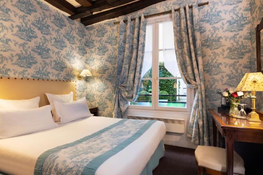 Cosy Parisian hotel room with blue and white toile de Jouy wallpaper, curtains and bed runner in matching fabric, exposed dark wooden beams, and a garden view window