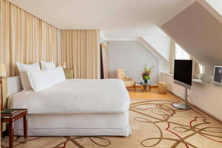 A spacious Paris hotel room with sloped ceilings, cream curtains, white bedding and an ornate patterned carpet