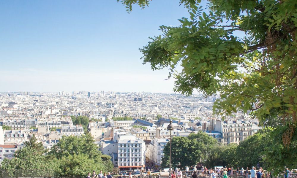 This is an image over an expansive view of Paris as a city, where you can see people below standing on a ledge looking out into the concrete jungle of buildings.