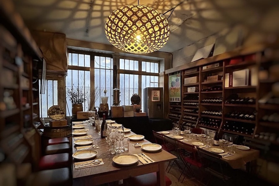 Rustic interior of a Paris wine bar set for a group dinner, featuring a long wooden table with place settings, floor-to-ceiling wine racks, and a decorative woven globe pendant light