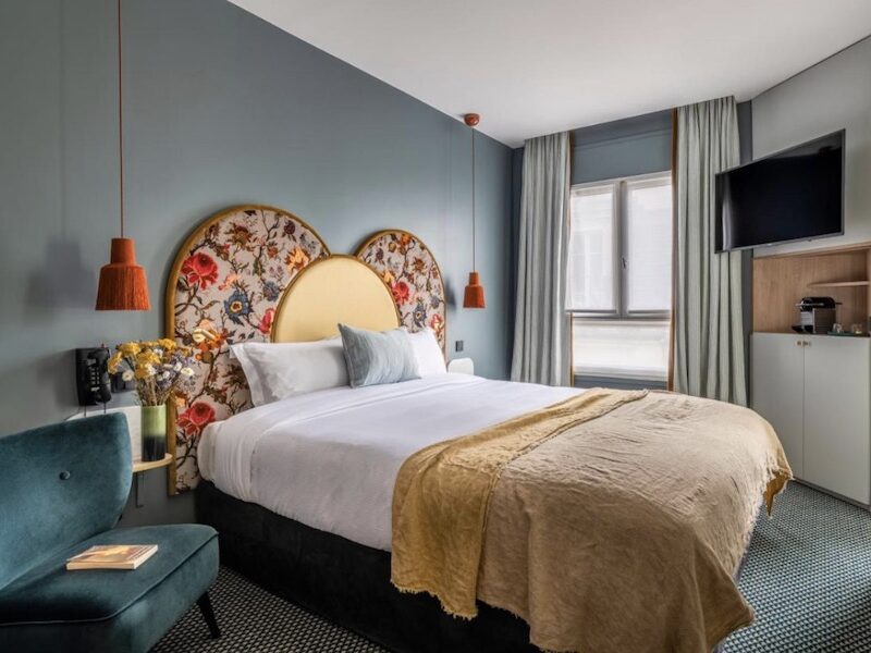 This is an image of a hotel room with blue painted walls. The bed is a double with a colourful headboard. There is a blue chair in the corner.